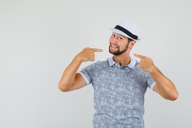 Varón joven apuntando a su sonrisa en camiseta, sombrero y mirando alegre, vista frontal.