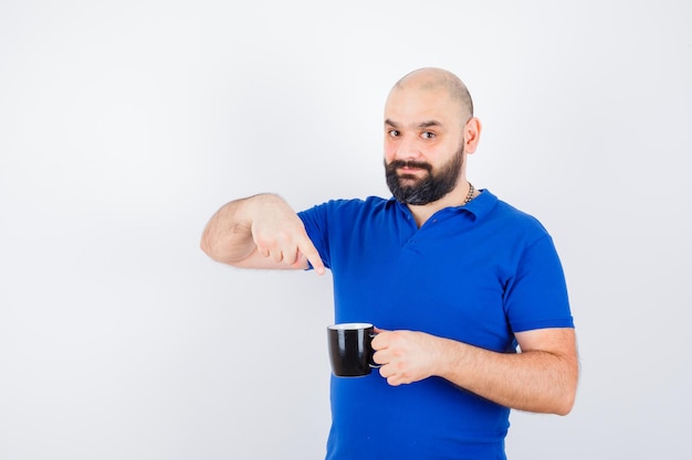 Varón joven apuntando a la taza negra con camisa azul y mirando positivo, vista frontal.