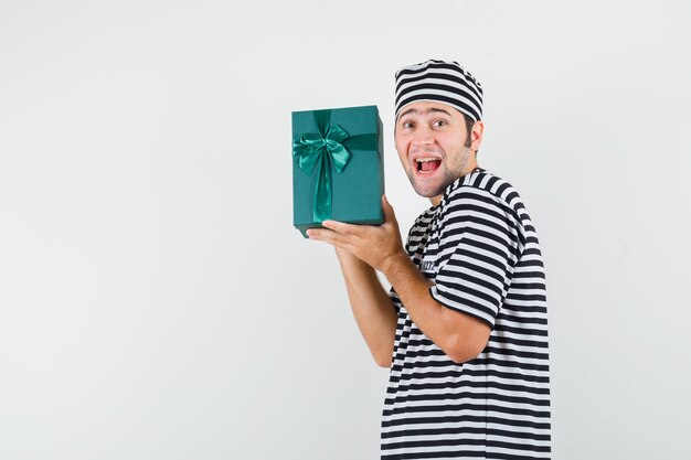 Varón joven con caja de regalo en camiseta, sombrero y mirando feliz.