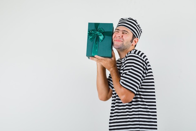 Varón joven con caja de regalo en camiseta, sombrero y mirando feliz.