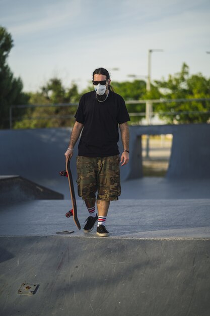 Varón joven caminando en un parque con una patineta con una mascarilla médica - Concepto Covid-19