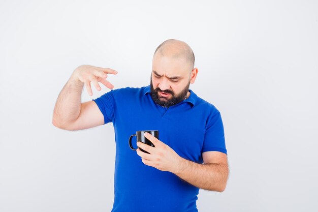 Varón joven con camisa azul mirando dentro de la taza mientras levanta la mano y mira insatisfecho, vista frontal.