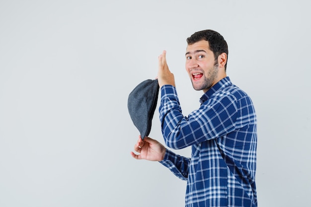 Varón joven con camisa azul mostrando gorra mientras mira a un lado y se ve alegre.