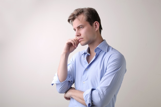 Varón joven con una camisa azul que tiene pensamientos profundos de pie contra una pared blanca