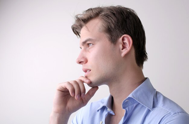 Varón joven con una camisa azul que tiene pensamientos profundos de pie sobre la pared blanca