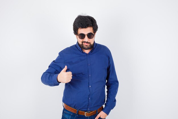 Varón joven con camisa azul real, gafas mostrando el pulgar hacia arriba y con un aspecto fresco, vista frontal.