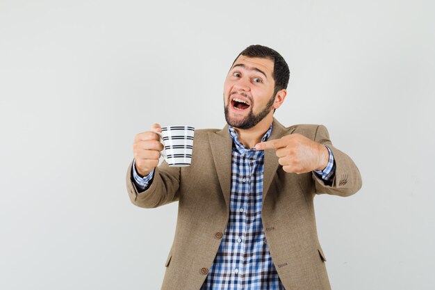 Varón joven en camisa, chaqueta apuntando a una taza de bebida y mirando contento, vista frontal.