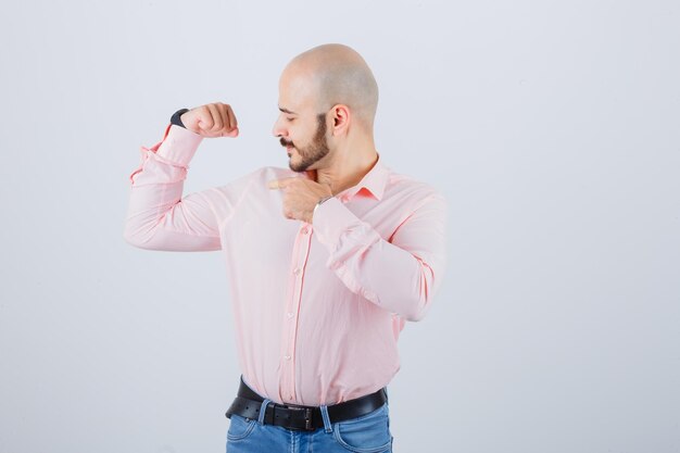 Varón joven en camisa, jeans apuntando los músculos del brazo y mirando orgulloso, vista frontal.