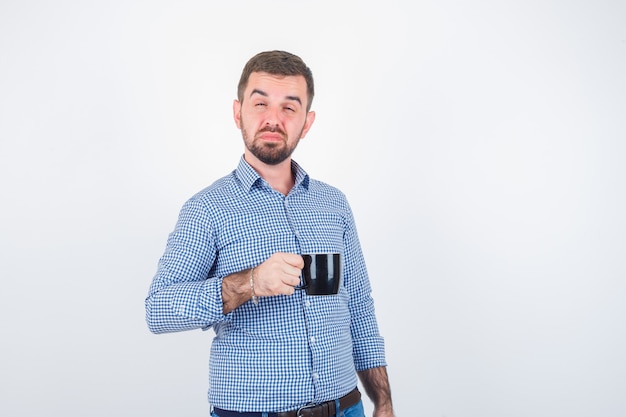 Varón joven en camisa, jeans cerrando los ojos mientras sostiene la taza y mirando confiado, vista frontal.