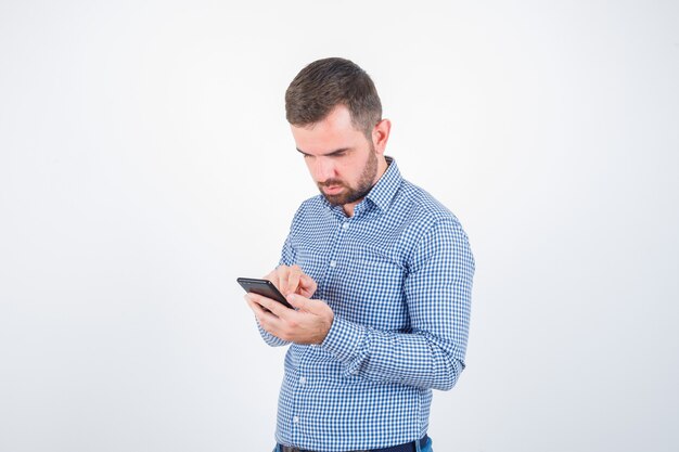 Varón joven en camisa, jeans mirando el teléfono móvil y mirando serio, vista frontal.