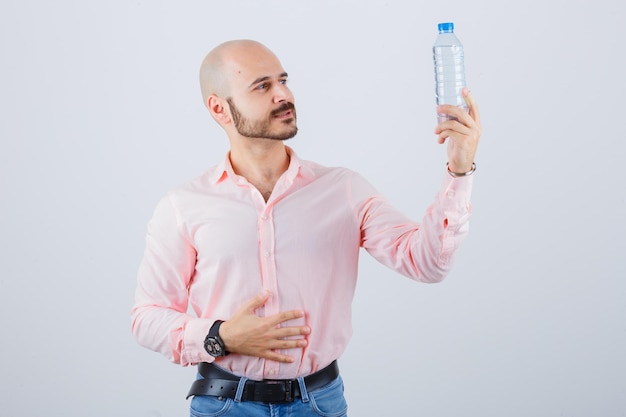 Varón joven en camisa, jeans sosteniendo una botella de agua de plástico y mirando enfocado, vista frontal.