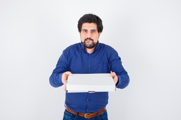 Varón joven en camisa, jeans sosteniendo una caja de pizza cerrada y mirando hambriento, vista frontal.