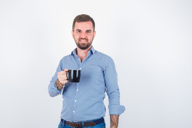 Varón joven en camisa, jeans sosteniendo la taza mientras está de pie y mirando alegre, vista frontal.