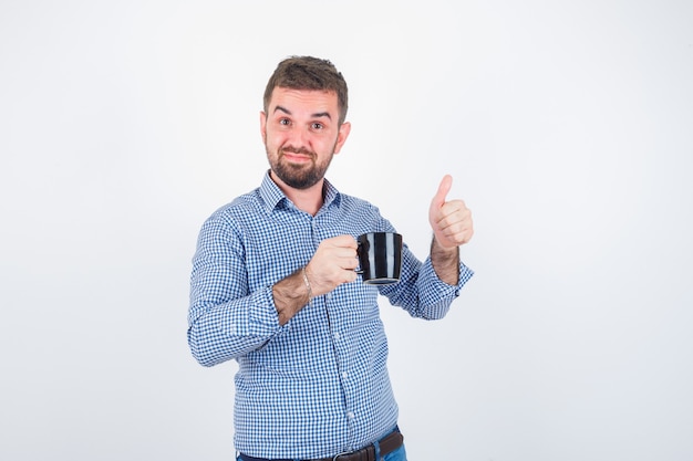 Varón joven en camisa, jeans sosteniendo la taza mientras muestra el pulgar hacia arriba y se ve feliz, vista frontal.