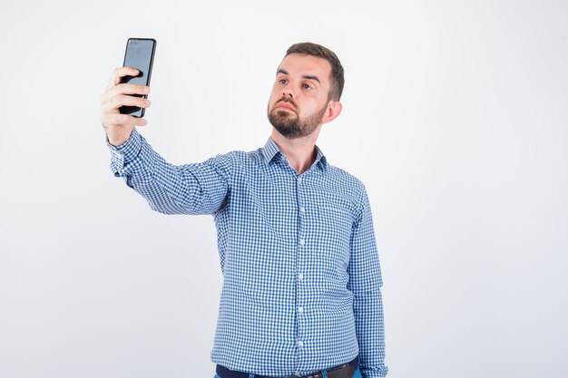 Varón joven en camisa, jeans tomando un selfie y luciendo guapo, vista frontal.