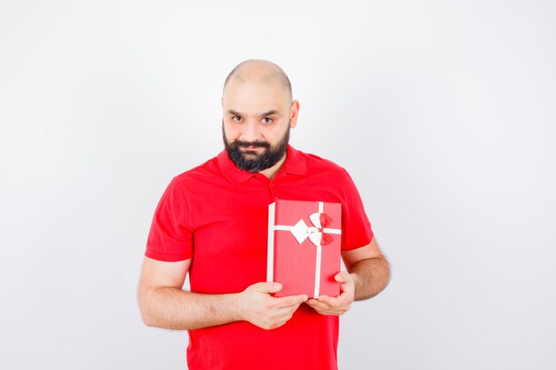 Varón joven en camisa roja mostrando caja de regalo y mirando complacido, vista frontal.