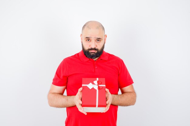 Varón joven en camisa roja que muestra la caja de regalo, vista frontal.