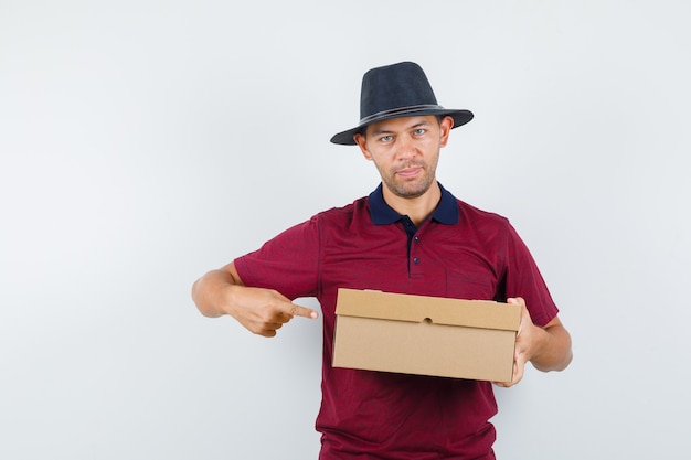 Varón joven en camisa roja, sombrero negro apuntando a cajas y mirando interesado, vista frontal.