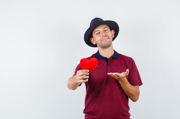 Varón joven en camisa roja, sombrero negro mostrando corazón rojo y mirando relajado, vista frontal.