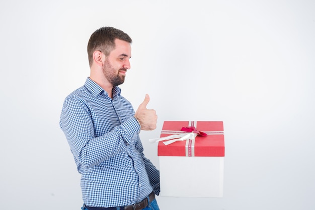 Varón joven en camisa sosteniendo una caja de regalo mientras muestra el pulgar hacia arriba y parece complacido, vista frontal