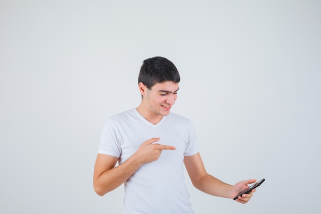 Varón joven en camiseta apuntando al teléfono y mirando alegre, vista frontal.