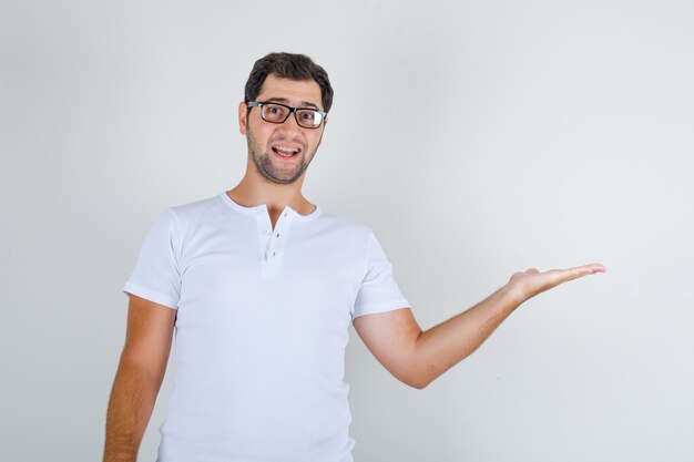 Varón joven en camiseta blanca, gafas mostrando algo o dando la bienvenida y mirando alegre