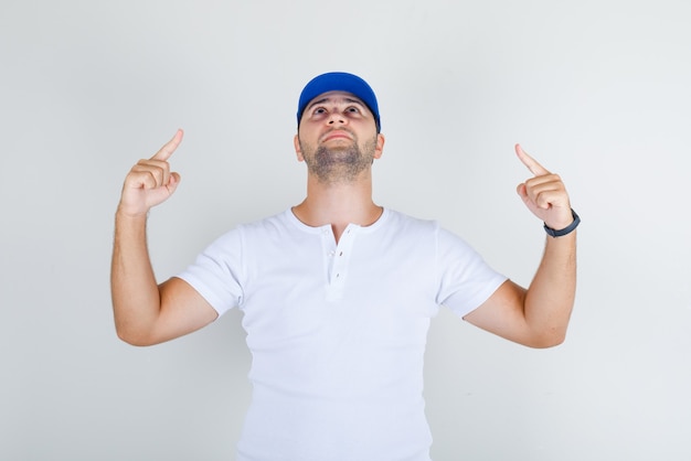 Varón joven en camiseta blanca, gorra azul apuntando hacia arriba con los dedos