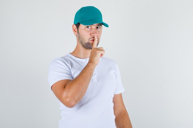 Varón joven en camiseta blanca con gorra haciendo gesto de silencio