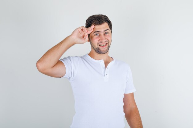 Varón joven en camiseta blanca manteniendo dos dedos en las sienes y mirando alegre