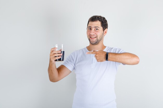 Varón joven en camiseta blanca mostrando bebida cola con el dedo y mirando feliz