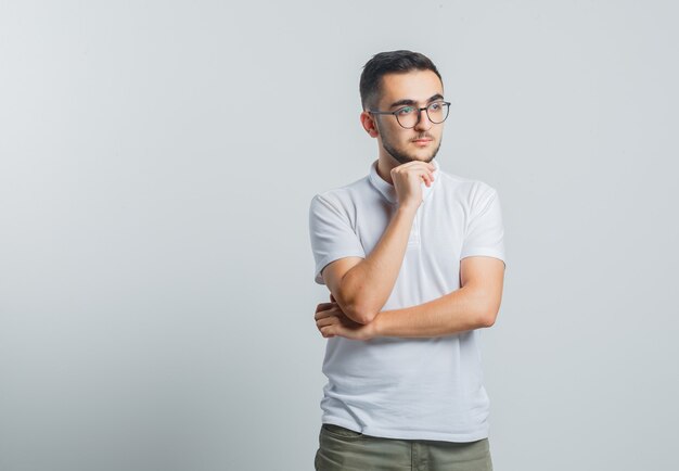 Varón joven en camiseta blanca, pantalones apoyando la barbilla en la mano y mirando pensativo