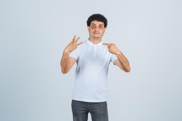 Varón joven en camiseta blanca, pantalones apuntando al número tres y mirando confiado, vista frontal.