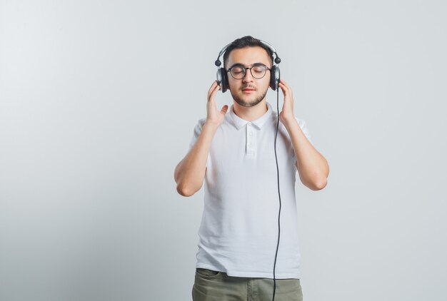 Varón joven en camiseta blanca, pantalones disfrutando de la música con auriculares y mirando relajado