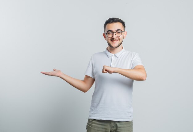 Varón joven en camiseta blanca, pantalones fingiendo tener reloj, extendiendo la palma a un lado y mirando confiado