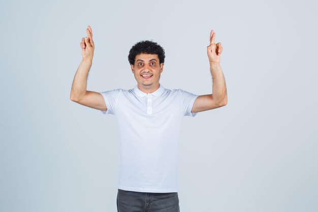 Varón joven en camiseta blanca, pantalones manteniendo los dedos cruzados y mirando feliz, vista frontal.