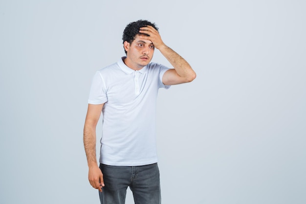 Varón joven en camiseta blanca, pantalones manteniendo la mano en la cabeza y mirando indefenso, vista frontal.