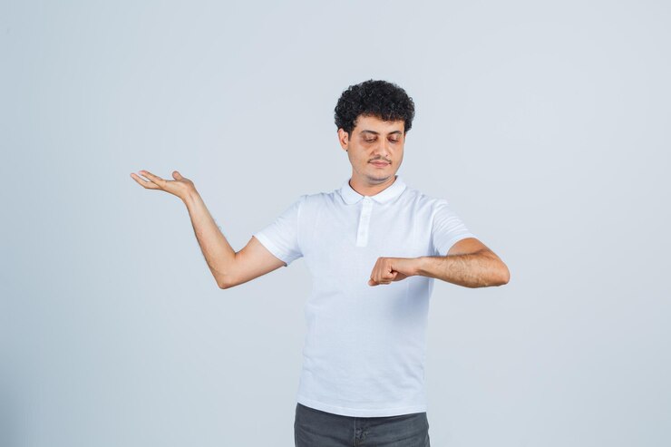 Foto gratuito varón joven en camiseta blanca, pantalones mirando el reloj fingió estar usado y luciendo confiado, vista frontal.