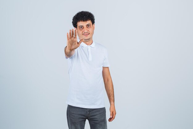 Varón joven en camiseta blanca, pantalones mostrando gesto de parada y mirando confiado, vista frontal.