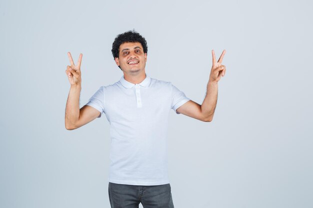 Varón joven en camiseta blanca, pantalones mostrando gesto de victoria y mirando confiado, vista frontal.