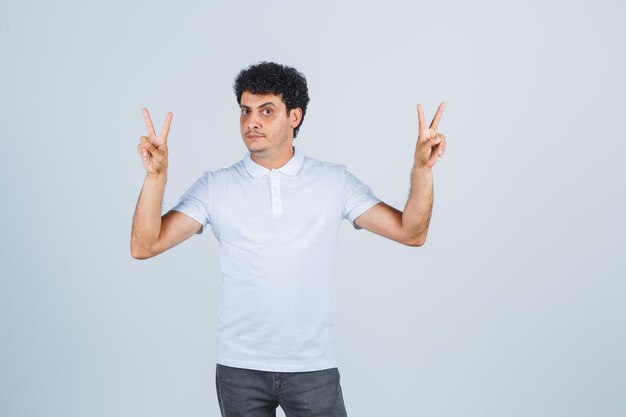 Varón joven en camiseta blanca, pantalones mostrando gesto de victoria y mirando confiado, vista frontal.