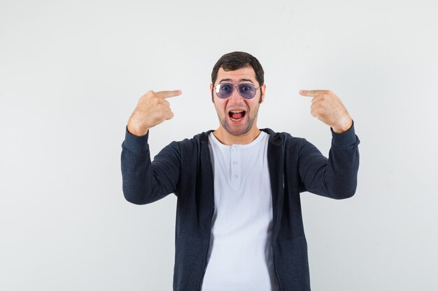 Varón joven en camiseta, chaqueta apuntando a sus gafas y mirando alegre, vista frontal.