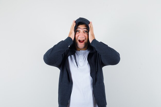 Varón joven en camiseta, chaqueta cogidos de la mano en la cabeza y mirando feliz, vista frontal.