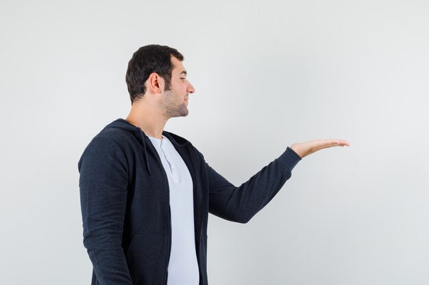 Varón joven en camiseta, chaqueta fingiendo sostener algo y mirando enfocado, vista frontal.