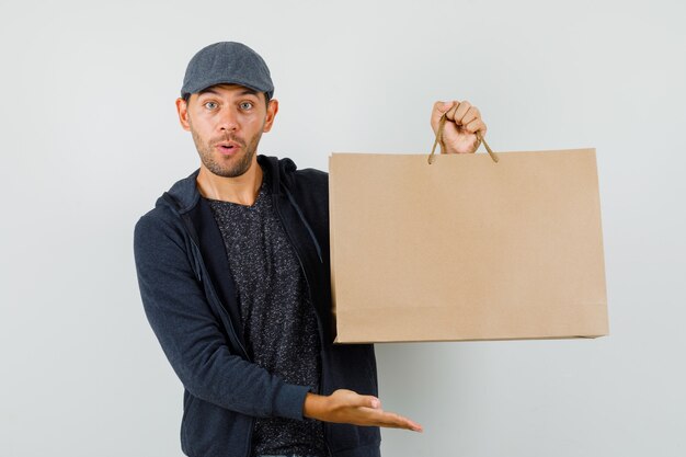 Varón joven en camiseta, chaqueta, gorra mostrando bolsa de papel y mirando asombrado, vista frontal.