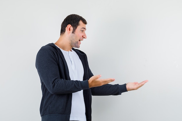 Varón joven en camiseta, chaqueta mostrando algo o dando la bienvenida y mirando alegre, vista frontal.