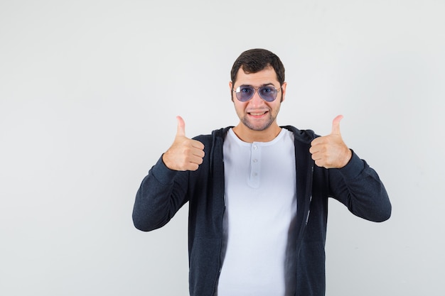 Varón joven en camiseta, chaqueta mostrando doble pulgar hacia arriba y mirando alegre, vista frontal.