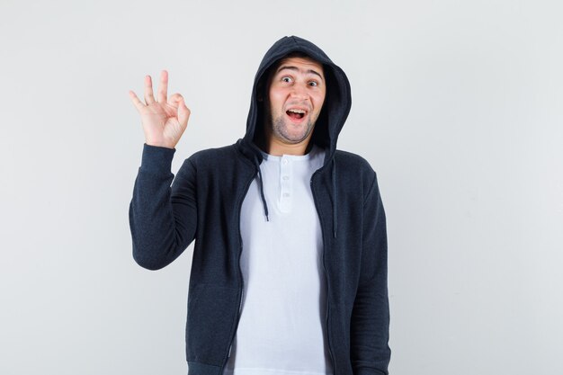 Varón joven en camiseta, chaqueta mostrando gesto ok y mirando alegre, vista frontal.