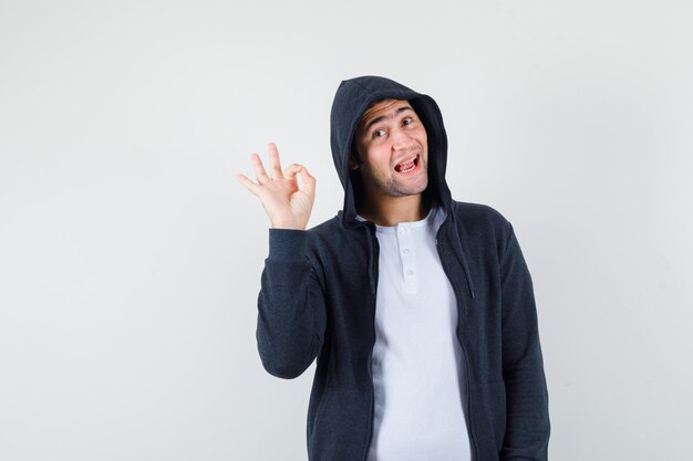 Varón joven en camiseta, chaqueta mostrando gesto ok y mirando alegre, vista frontal.