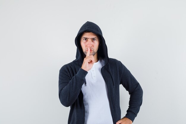 Varón joven en camiseta, chaqueta mostrando gesto de silencio y mirando serio, vista frontal.