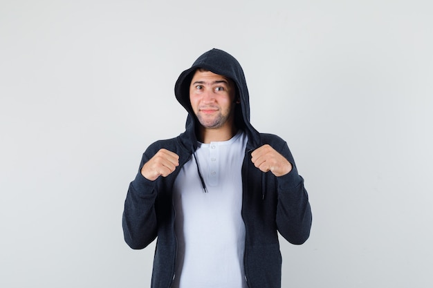 Varón joven en camiseta, chaqueta de pie en pose de lucha y mirando confiado, vista frontal.